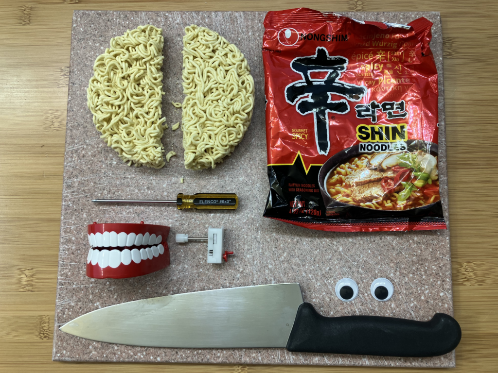 Photo of several incongruous items arranged neatly on a cutting board: a large kitchen knife, a packet of ramen noodles, a round of dried ramen boodles that has been cut in two, a small screwdriver, 2 medium-size googly eyes, a life-size plastic chatter teeth with the chatter mechanism removed and laid next to it