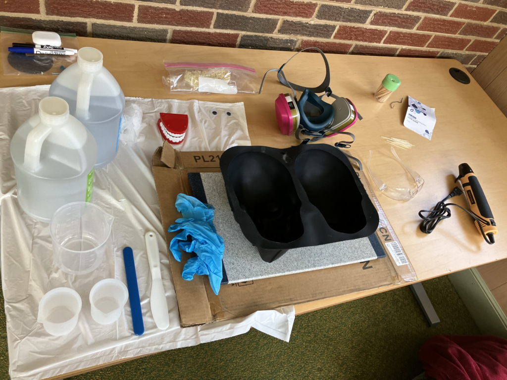 Photo of a desk laid out with various tools and supplies for resin casting, including a life-size skull silicone mold, two big jugs of resin, measuring cups, safety mask, and a heat gun