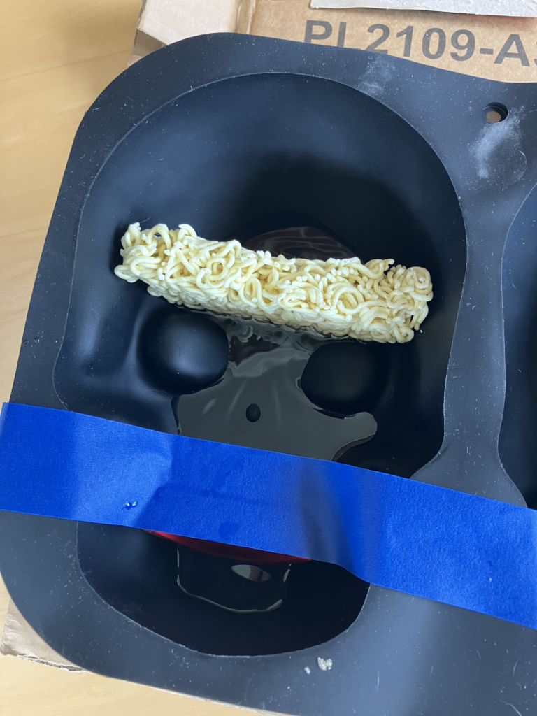 Photo of a black silicone full-size-skull cake mold. You can see half a cake of dried ramen noodles lodged in the skull's brain area, and blue painter's tape holding chatter teeth in place in the skull's teeth area. There is a small amount of resin on the very bottom of the mold, less than 1" deep.