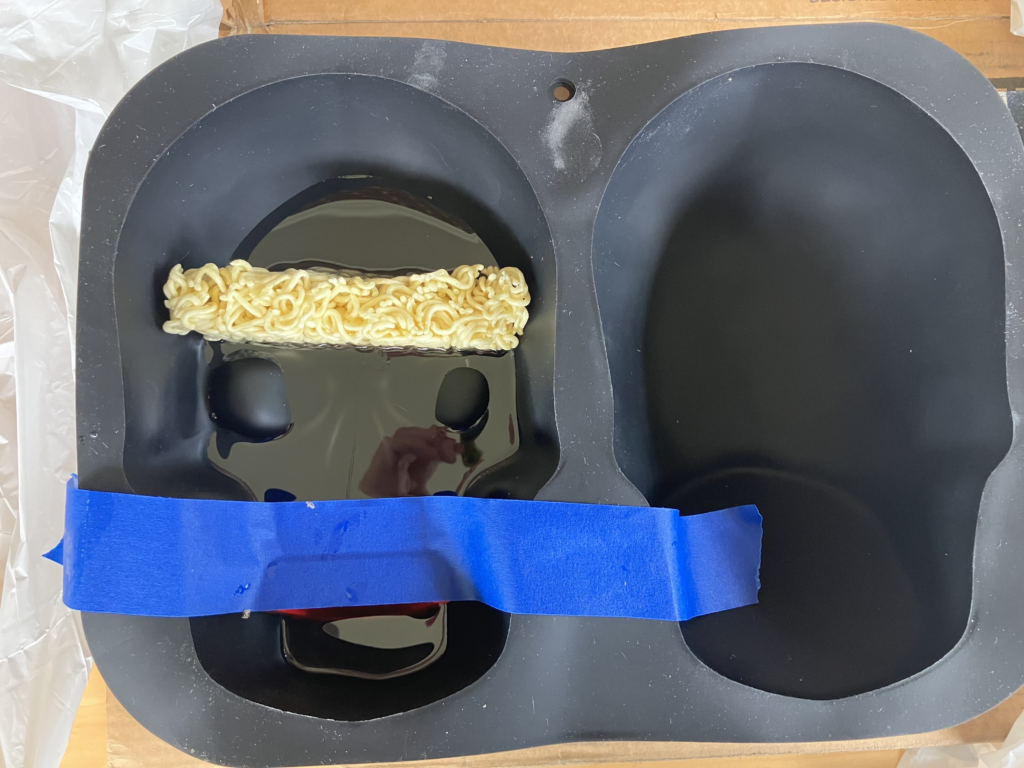 Photo of a black silicone full-size-skull cake mold. You can see half a cake of dried ramen noodles lodged in the skull's brain area, and blue painter's tape holding chatter teeth in place in the skull's teeth area. There is a small amount of resin on the very bottom of the mold, around 1" deep.