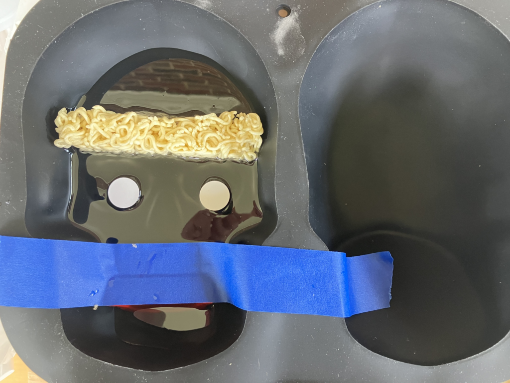 Photo of a black silicone full-size-skull cake mold. You can see half a cake of dried ramen noodles lodged in the skull's brain area, and blue painter's tape holding chatter teeth in place in the skull's teeth area. There is a small amount of wet resin on top of dried resin on the very bottom of the mold, around 1.5" deep. Two 1" googly eyes have been set upside-down into the resin over the skull's eye areas.