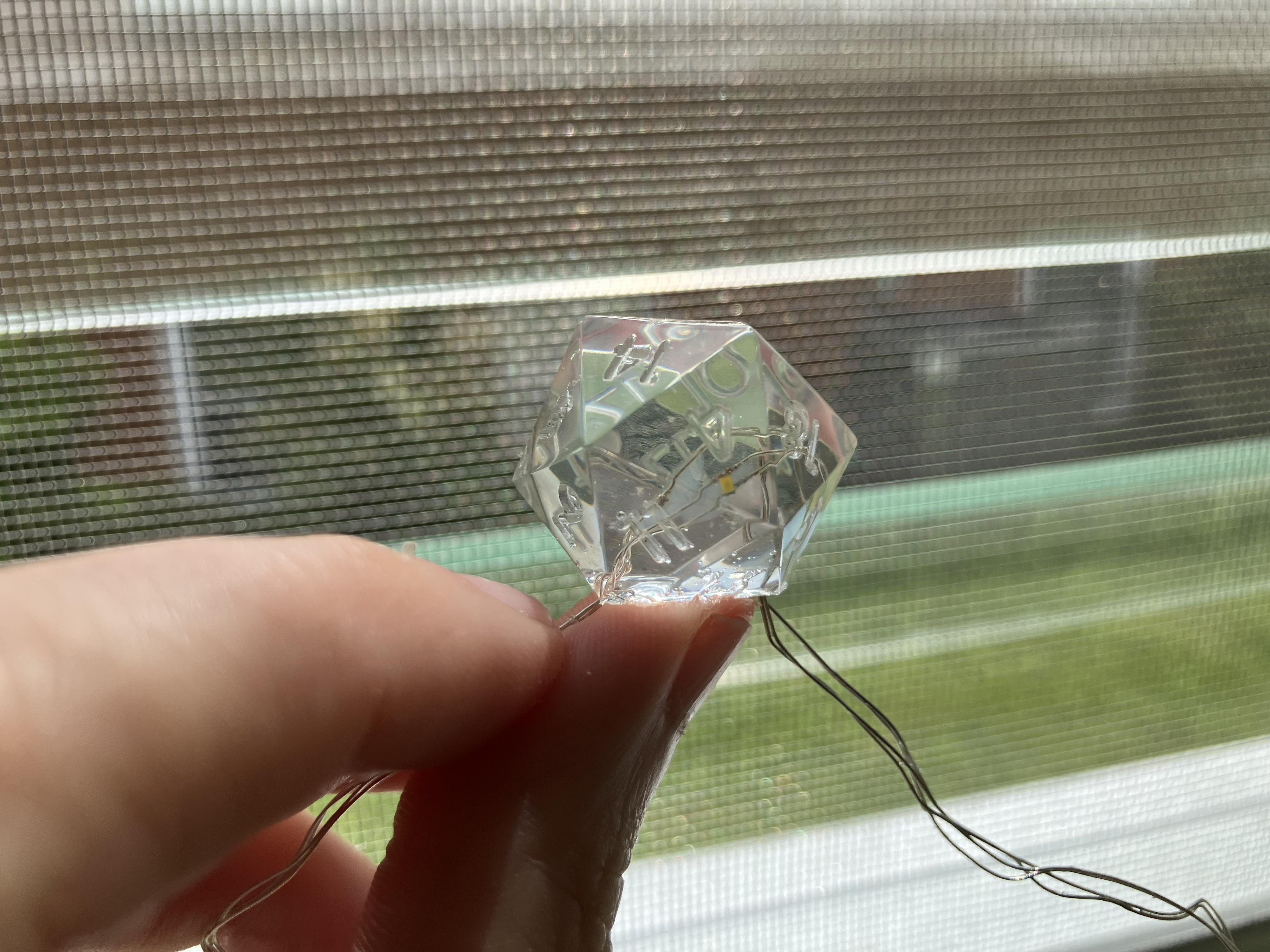 Photo of a transparent resin d20 die close up, with silver wires leading into it where an LED bulb is embedded inside the transparent resin