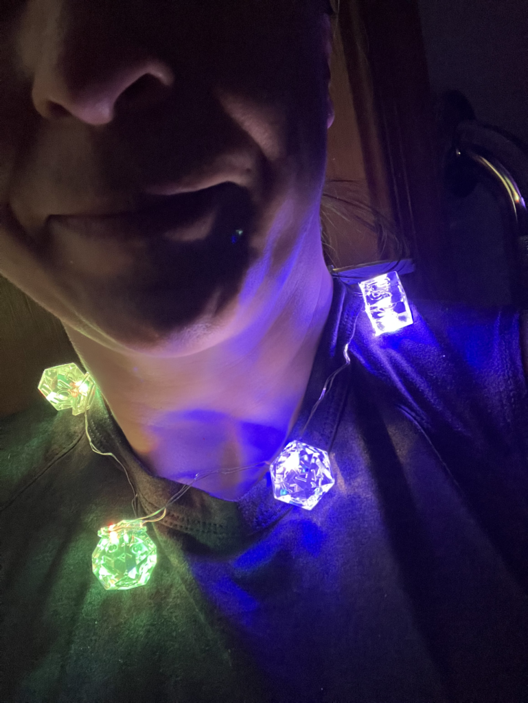 Photo of a person in a dark room, wearing a necklace of TTRPG transparent resin dice lit up from inside with LEDs in various colors