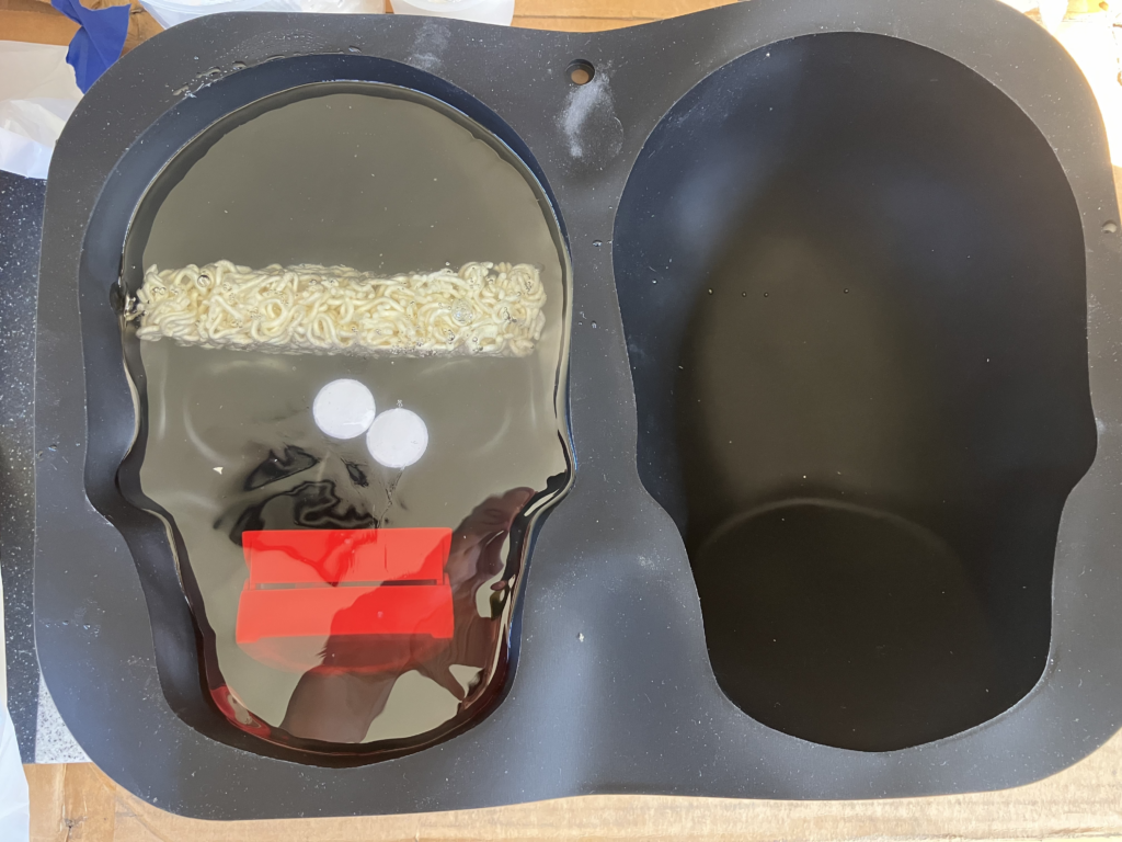Photo of a black silicone full-size-skull cake mold. You can see half a cake of dried ramen noodles lodged in the skull's brain area, chatter teeth in the skull's teeth area, and upside down googly eyes in the center but not correctly over the eye sockets. There is a of set resin on the very bottom of the mold, around 3" deep. The chatter teeth, googly eyes, and ramen noodles are now all fully encased/embedded inside the transparent resin.