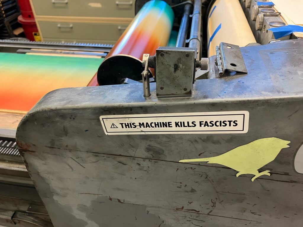 Photo of the side of a large metal printing press, with a "this machine kills fascists" sticker on it. Visible above are the printer rollers and bed, which are both colored with a gradient of ink from red to pale orange to green.