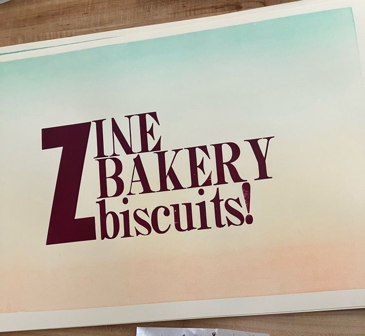 Photo of the words "Zine Bakery biscuits!" letterpress-printed in crimson red, on a background of gradient-colored green-yellow-orange paper.