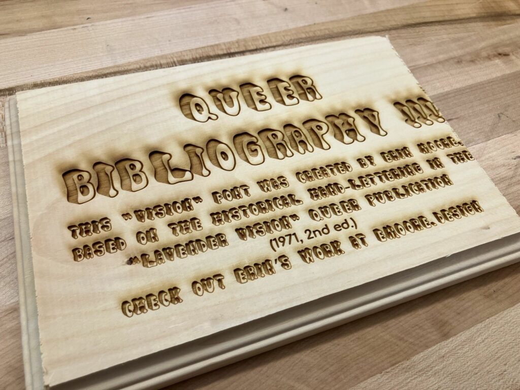 Lasercut wood that says "queer bibliography " and explains font was made by Erin Moore from a 1971 handlettered publication